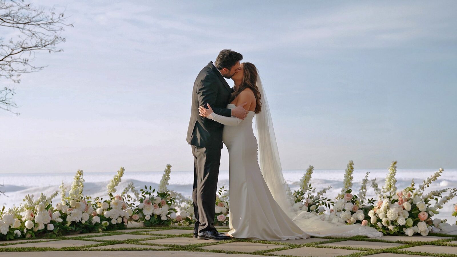 Still frame of a bride and groom kissing during their Lake Michigan wedding ceremony at Lighthouse Bluff Airbnb, surrounded by white and blush florals.