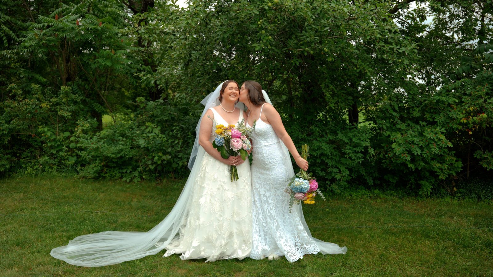 Newlyweds posing together on the lawn with one kissing the other on the cheek.